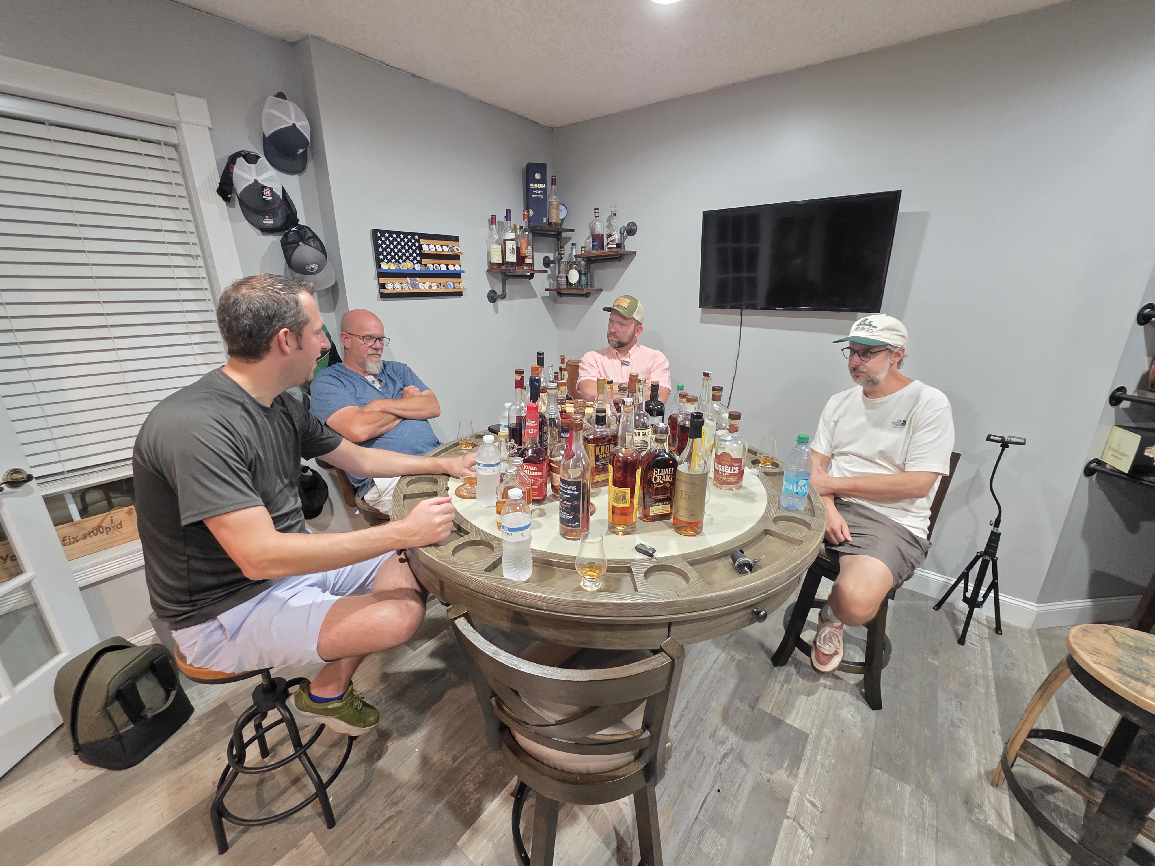 Four friends seated around a barrel table covered with bourbon bottles late in the evening.